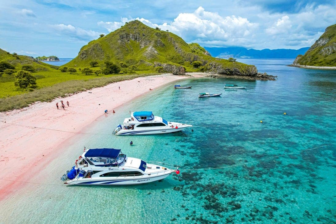 Kapal bersandar di pantai berpasir merah muda di bawah bukit hijau